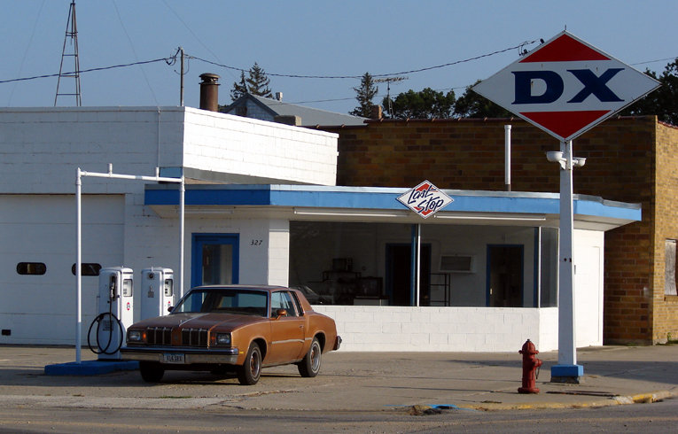Palmer, IA: An Oldsmobile pauses for a top-off at the "Last Stop" DX station, yet, the hapless driver will have to wait far longer than expected. The station ceased operating twenty years ago, and today stands proudly as a well-preserved museum piece, beloved by the town.