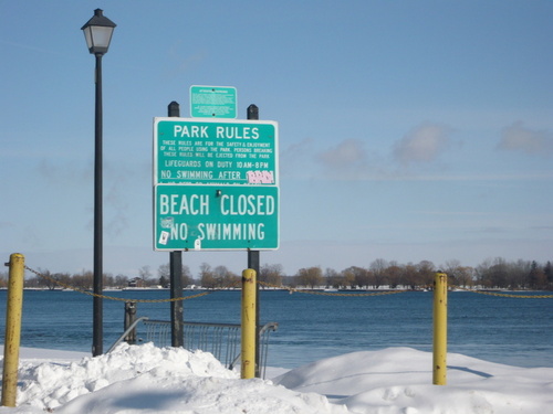 Marine City, MI: Beach is closed No swimming! sign with a light pole next to it. The sun shining on top of the water. Snow covers most of the sand.Yellow poles that hold chains.